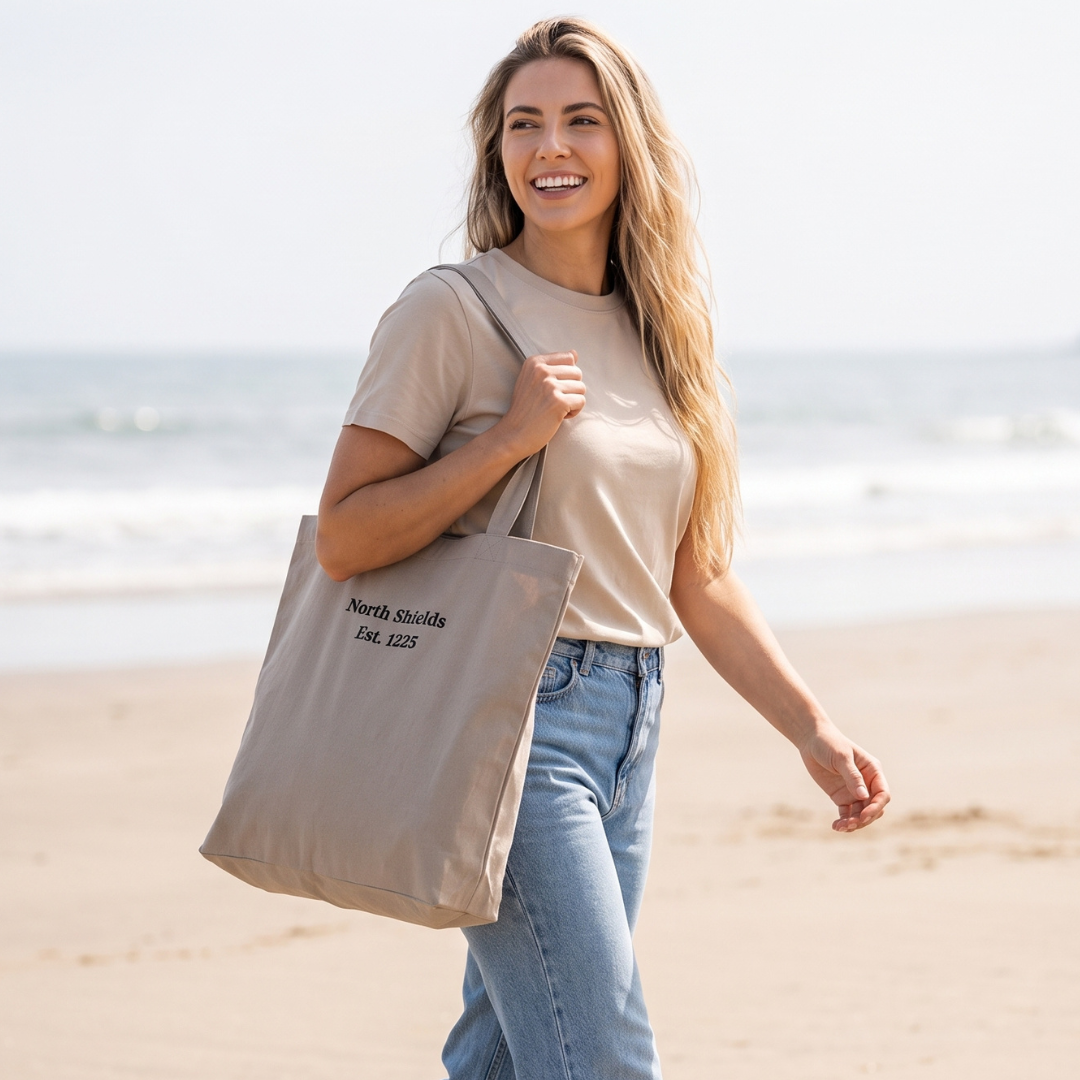 A woman walking on a sandy beach carrying a beige canvas tote bag with black printed text that reads 'North Shields Est. 1225'.