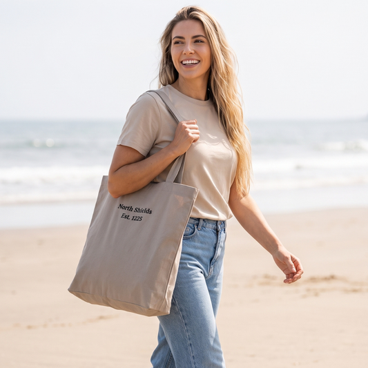 A woman walking on a sandy beach carrying a beige canvas tote bag with black printed text that reads 'North Shields Est. 1225'.