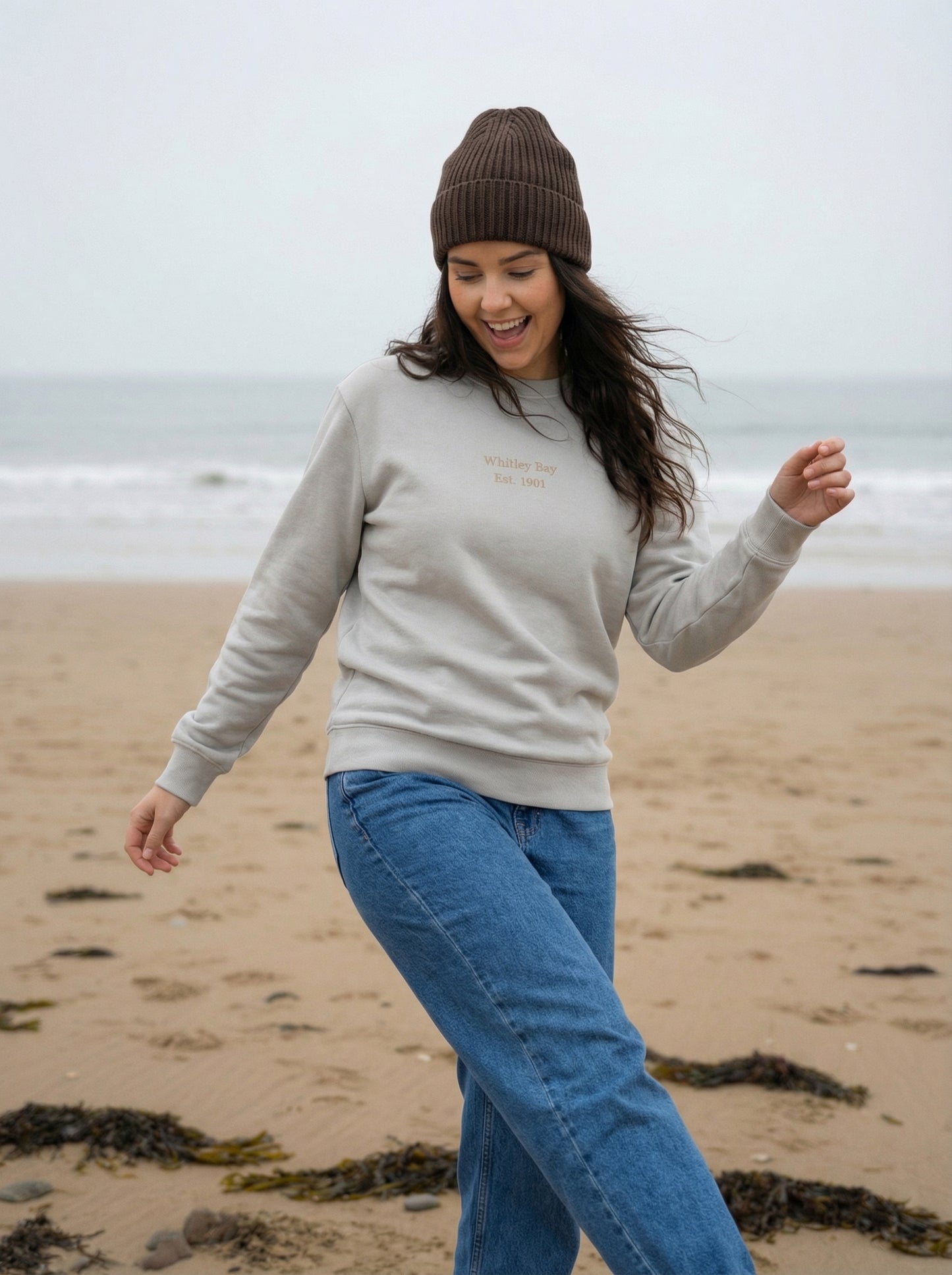 Woman in a gray sweatshirt and brown beanie walking on a beach.